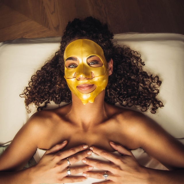 A woman at Neptune SPA, wearing a gold face mask is peacefully lying on a bed, indulging in a relaxing skincare routine.